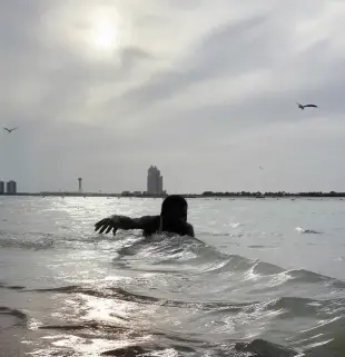 AI Video: The man in the water begins swimming freestyle toward the shore, moving his...